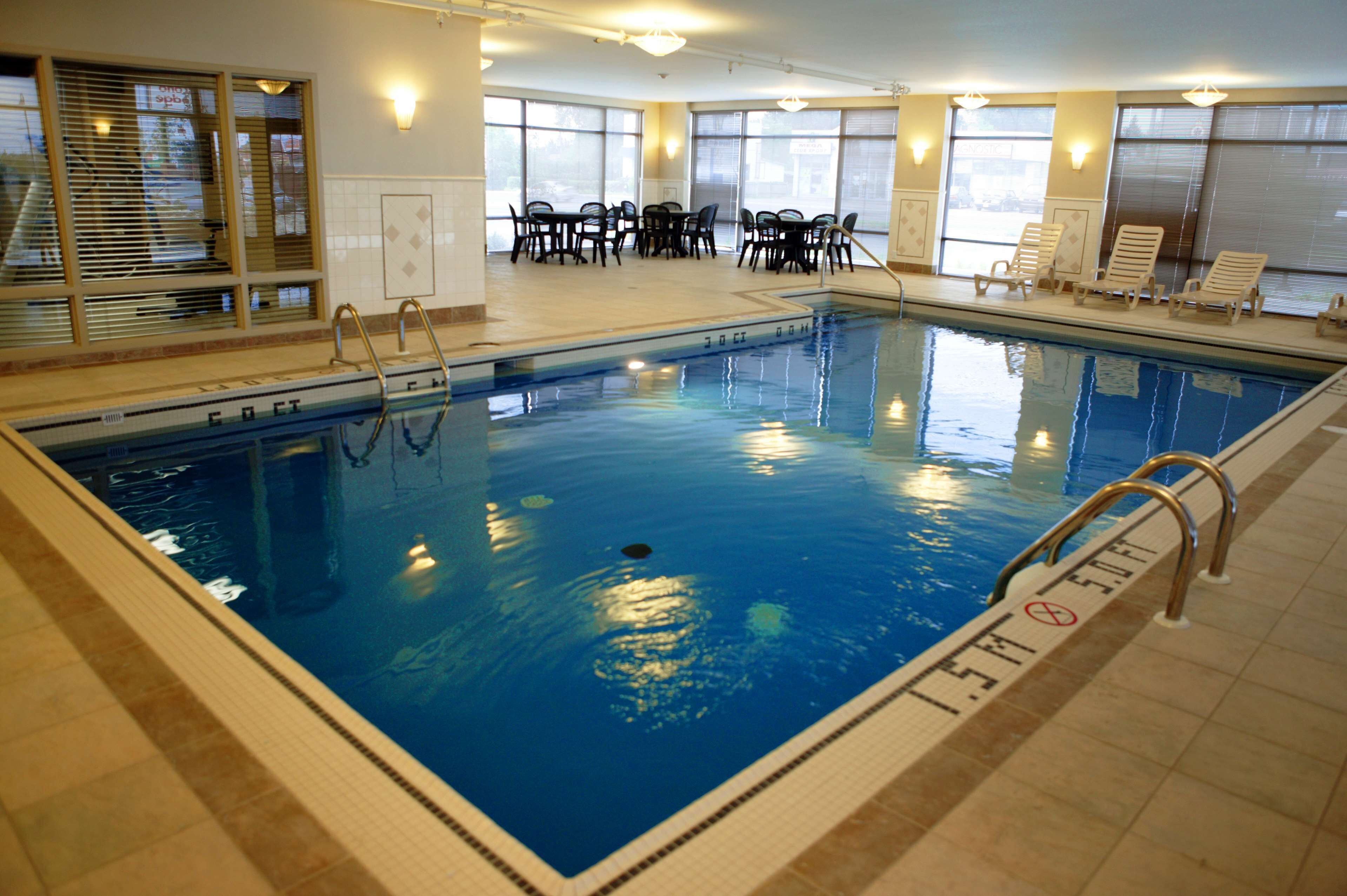 Una piscina interior con agua cristalina, una escalera de piscina y suelo de baldosas. Hay sillas y mesas alrededor de la zona de la piscina, y grandes ventanales aportan luz natural.