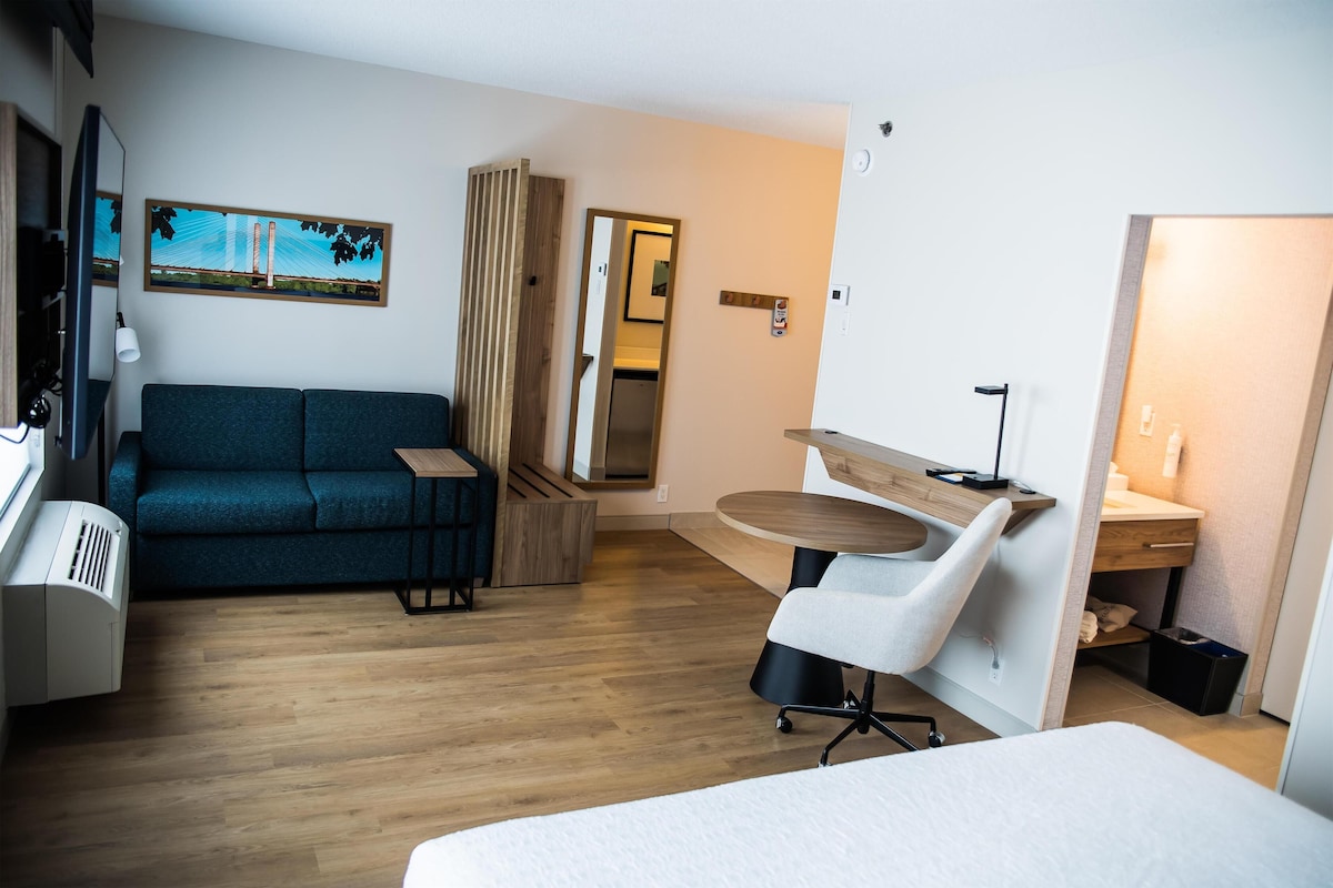 A modern hotel room with a sofa, a small round table, a chair, a bed, a desk, and a bathroom.
