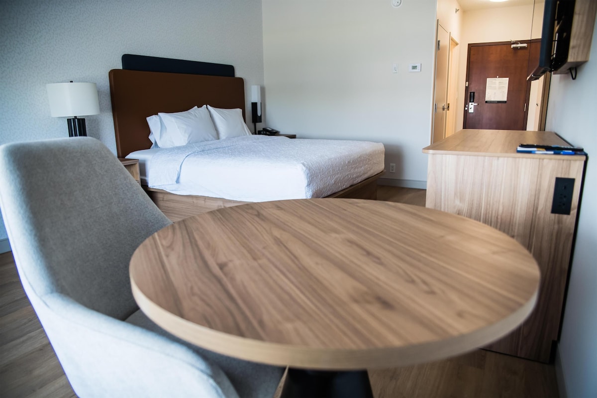 A hotel room with a bed, a bedside table, a chair, and a round table.