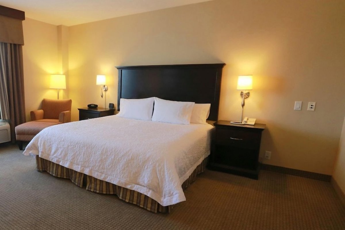 A hotel room with a large bed, two bedside tables with lamps, a chair, and a carpeted floor.