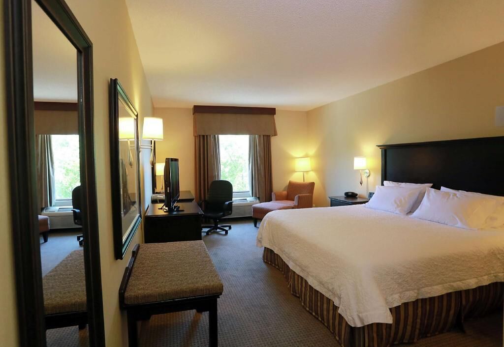 A hotel room with a large bed, a desk with a chair, a small table with a lamp, a chair, a mirror, and a window with curtains.