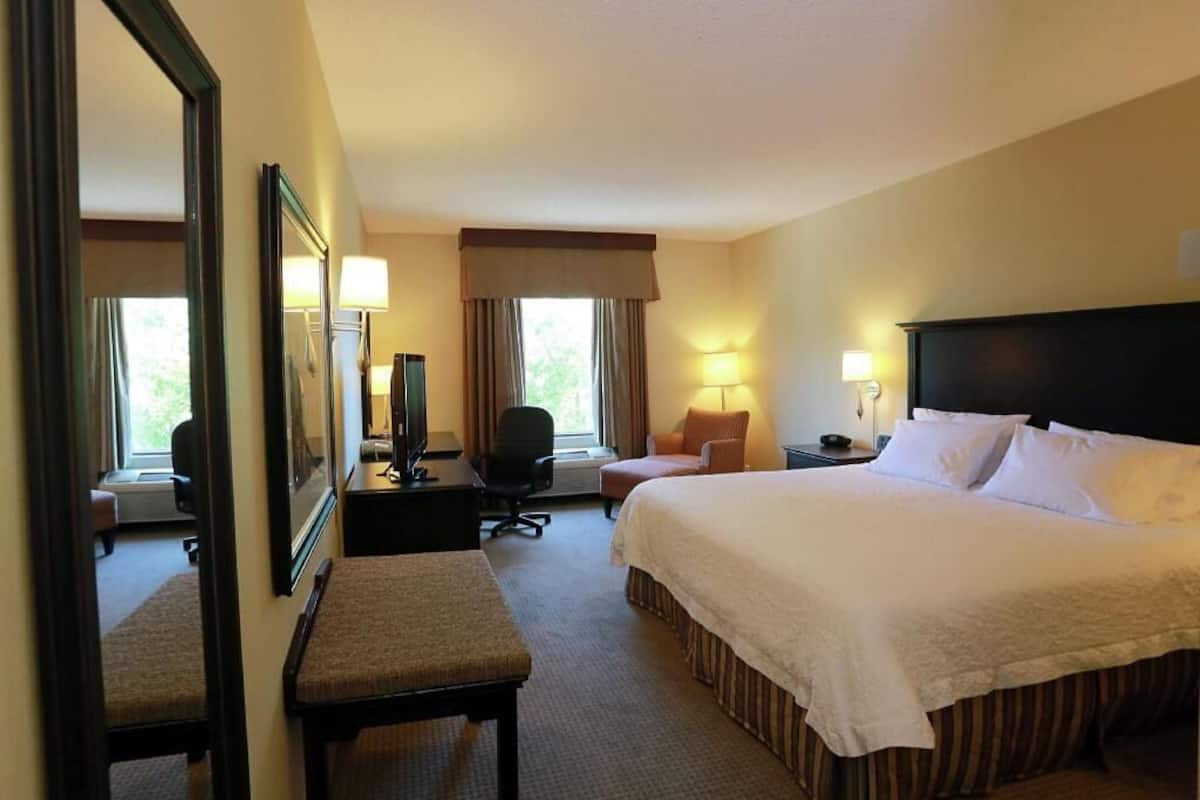 Habitación de hotel con una cama grande, un escritorio con silla, una mesita con lámpara, un sillón y un espejo, además de una ventana con cortinas.