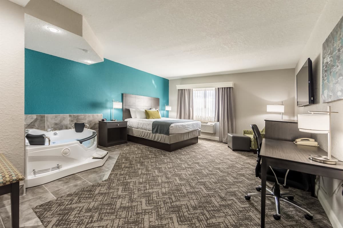 A modern hotel room with a large bed, a desk, a chair, a desk lamp, a flat-screen TV, a bathroom with a bathtub, and a window with curtains.