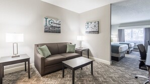 A hotel room with a brown leather sofa, a small table, two wall-mounted lamps, and a bed with a gray headboard. - Best Western Plus Sebastian Hotel & Suites (Sebastian)