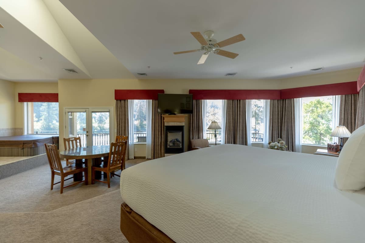 A hotel room with a large bed, a dining table with chairs, a fireplace, and a balcony with a view of trees.