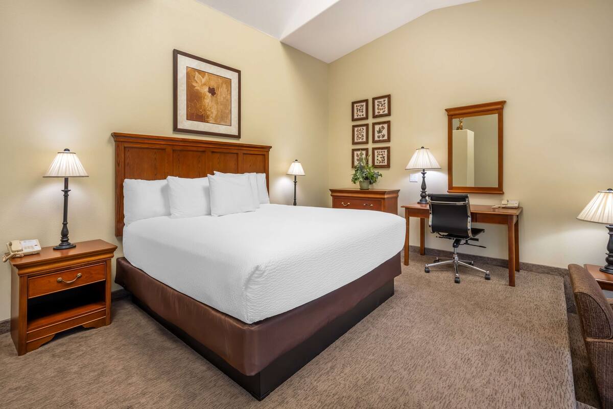 A hotel room with a large bed, bedside tables, a desk, a chair, a mirror, and framed artwork on the walls.