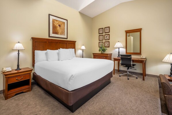A hotel room with a large bed, bedside tables, a desk, a chair, a mirror, and framed artwork on the walls.