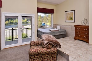 A room with a view of a river, a hot tub, a wooden dresser, and a chair with a blanket.