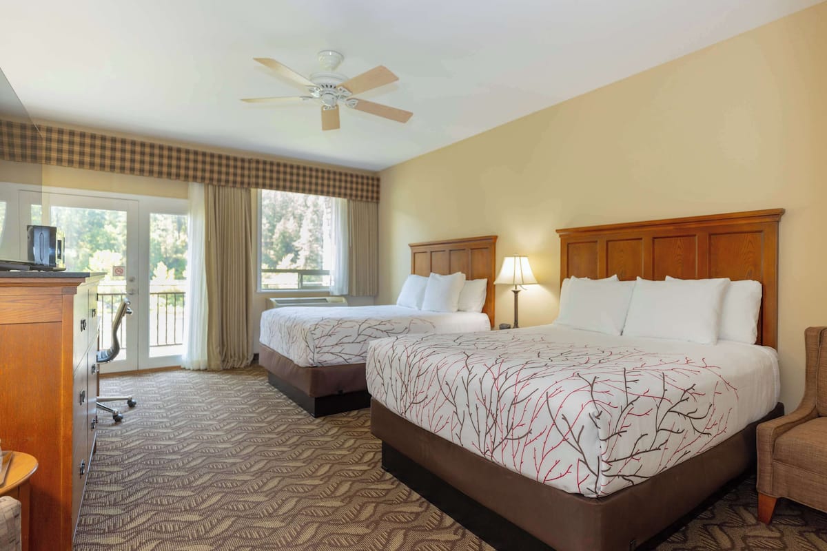 A hotel room with two beds, a ceiling fan, a desk with a chair, and a view of the outdoors.