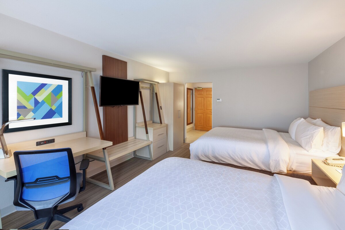 A hotel room with a desk, chair, TV, two beds, and a door leading to a bathroom.