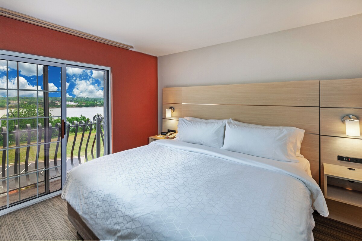A hotel room with a large bed, a nightstand, a lamp, and a view of a body of water through a sliding door.