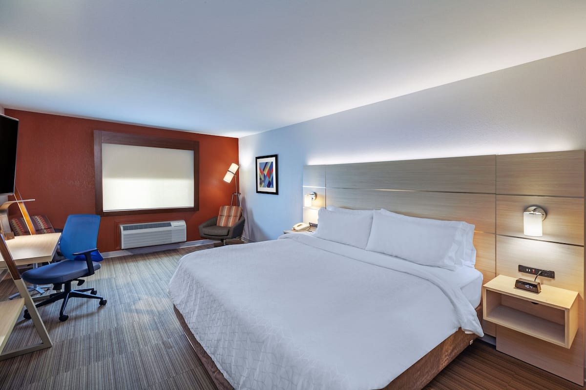 A hotel room with a large bed, a desk with a chair, a desk lamp, a wall-mounted phone, and a bedside table with a lamp.