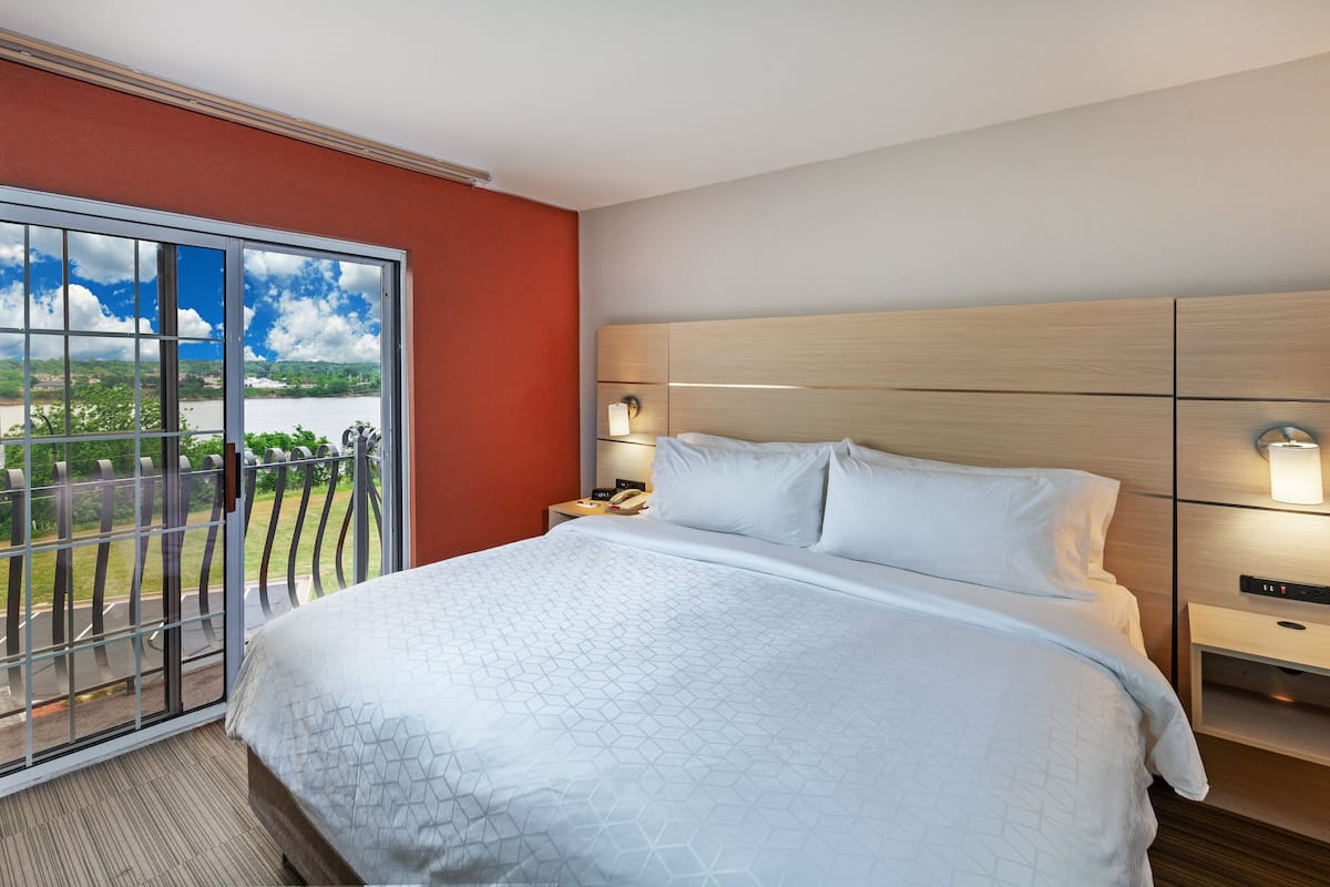 A hotel room with a large bed, a nightstand, a lamp, and a view of a balcony with a railing.