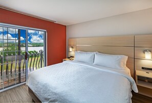 A hotel room with a large bed, a nightstand, a lamp, and a view of a balcony with a railing. - Holiday Inn Express Hotel & Suites Jenks by IHG (Jenks)