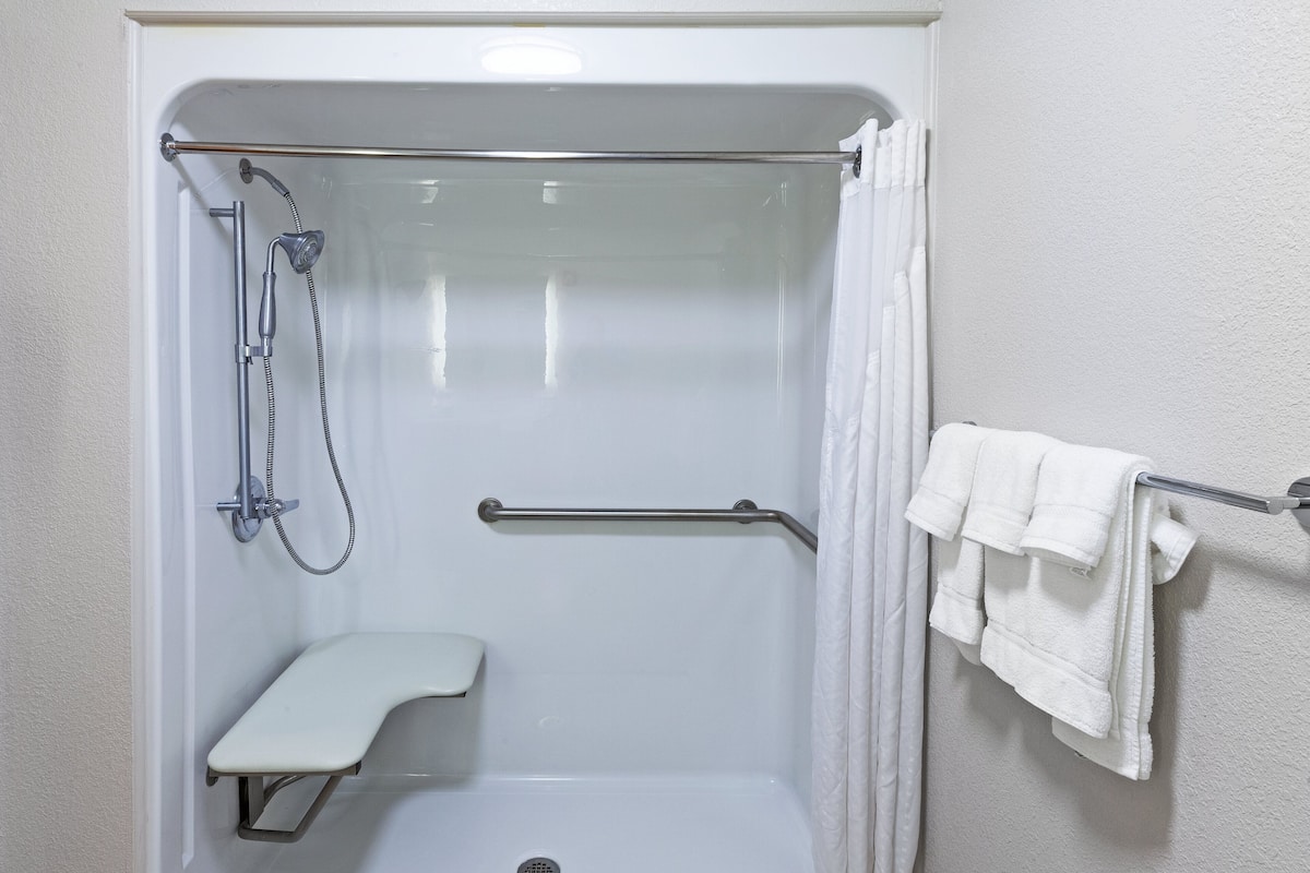 A white shower stall with a handheld showerhead, a grab bar, a bench, and a towel rack with towels.