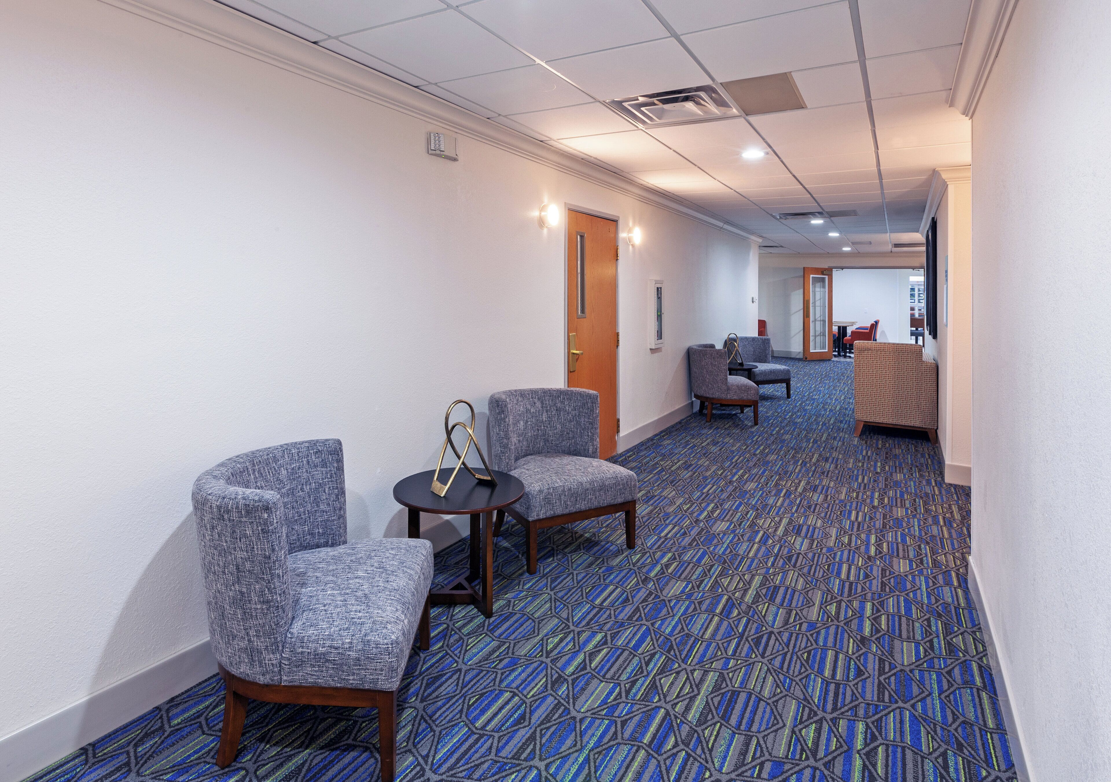 A hotel corridor with seating areas, a patterned carpet, and a door leading to another room.