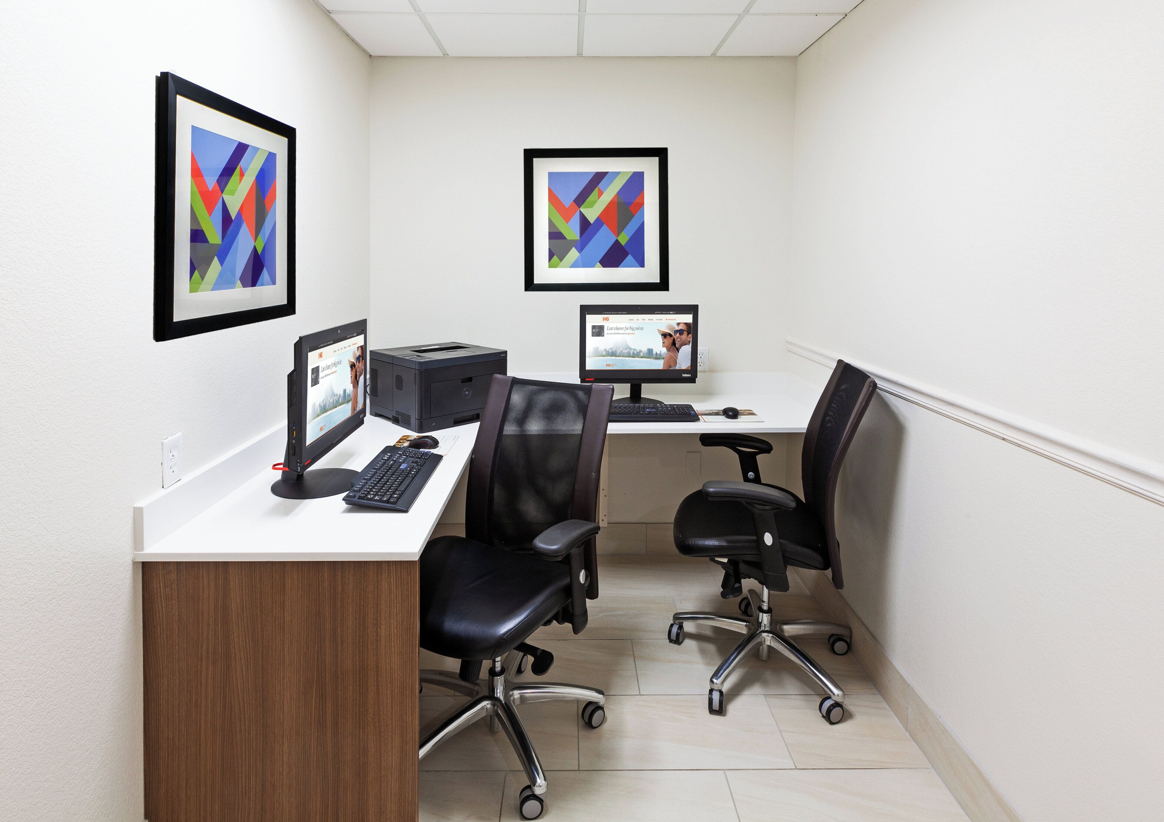 Un espace de bureau moderne comprenant deux bureaux, des chaises, des ordinateurs et des œuvres d’art abstraites encadrées accrochées aux murs.