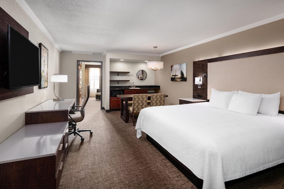 A hotel room with a large bed, a desk with a chair, a television, a desk lamp, a mirror, and a small kitchenette.