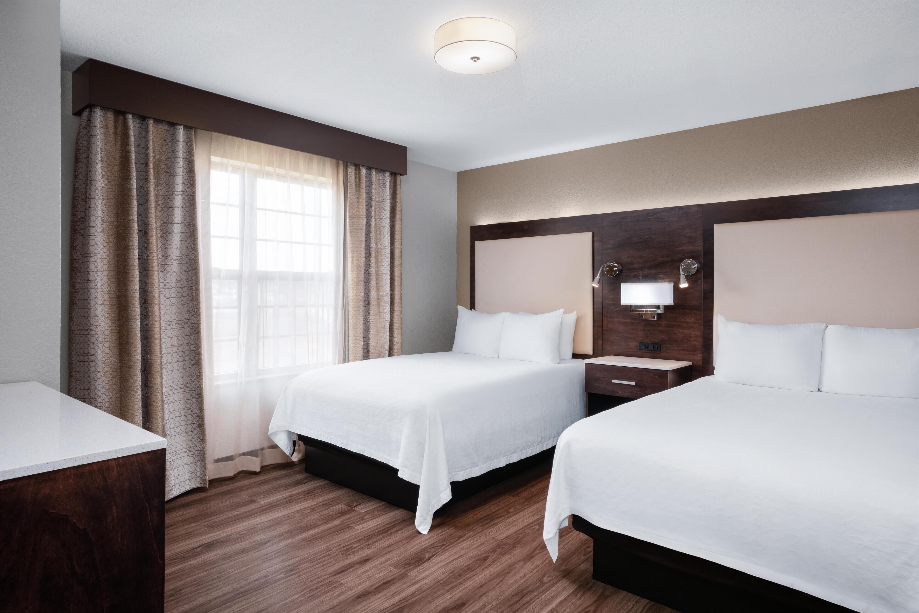 A hotel room with two beds, a wooden floor, a large window with curtains, and a wall-mounted light fixture.