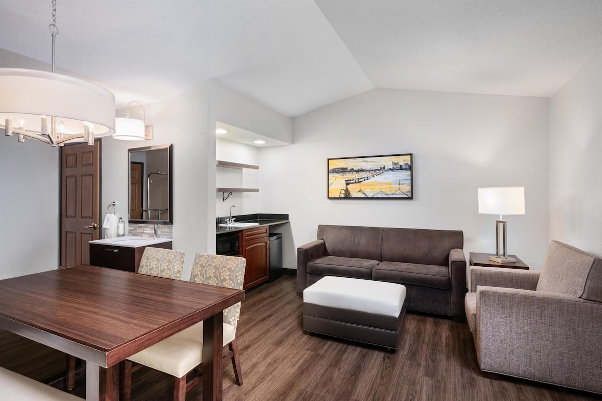 A modern hotel room with a dining area, kitchenette, and a living space with a sofa and armchairs.