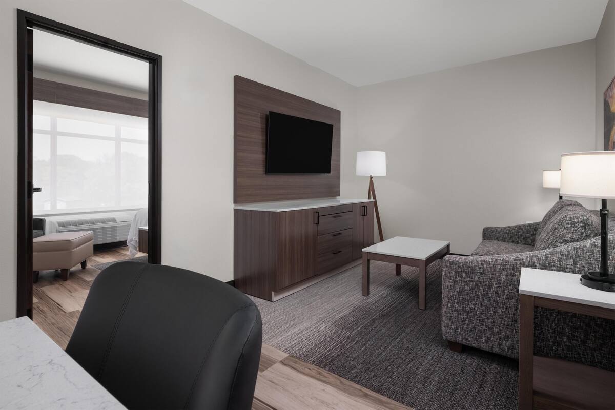 A modern hotel room with a flat-screen TV mounted on a wooden panel, a small table with a lamp, a sofa, and a chair.