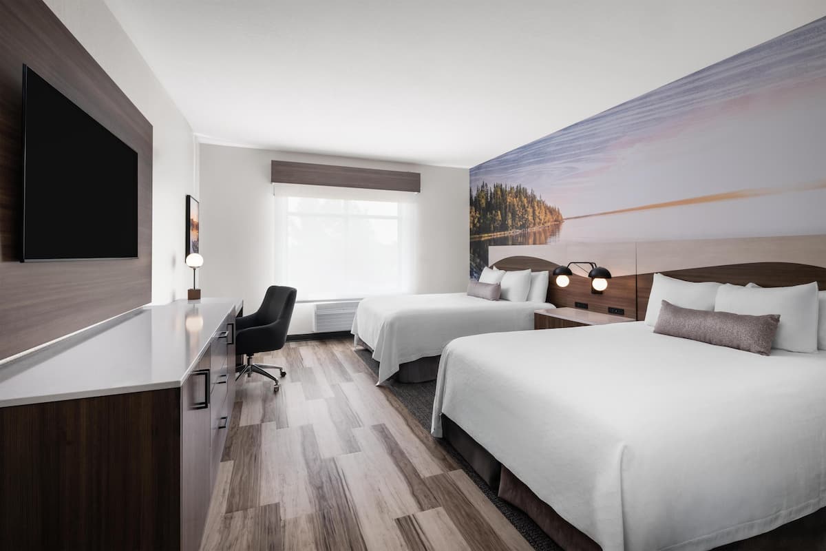 A hotel room with two beds, a desk, a chair, a flat-screen TV, and a large window with a view of a lake.