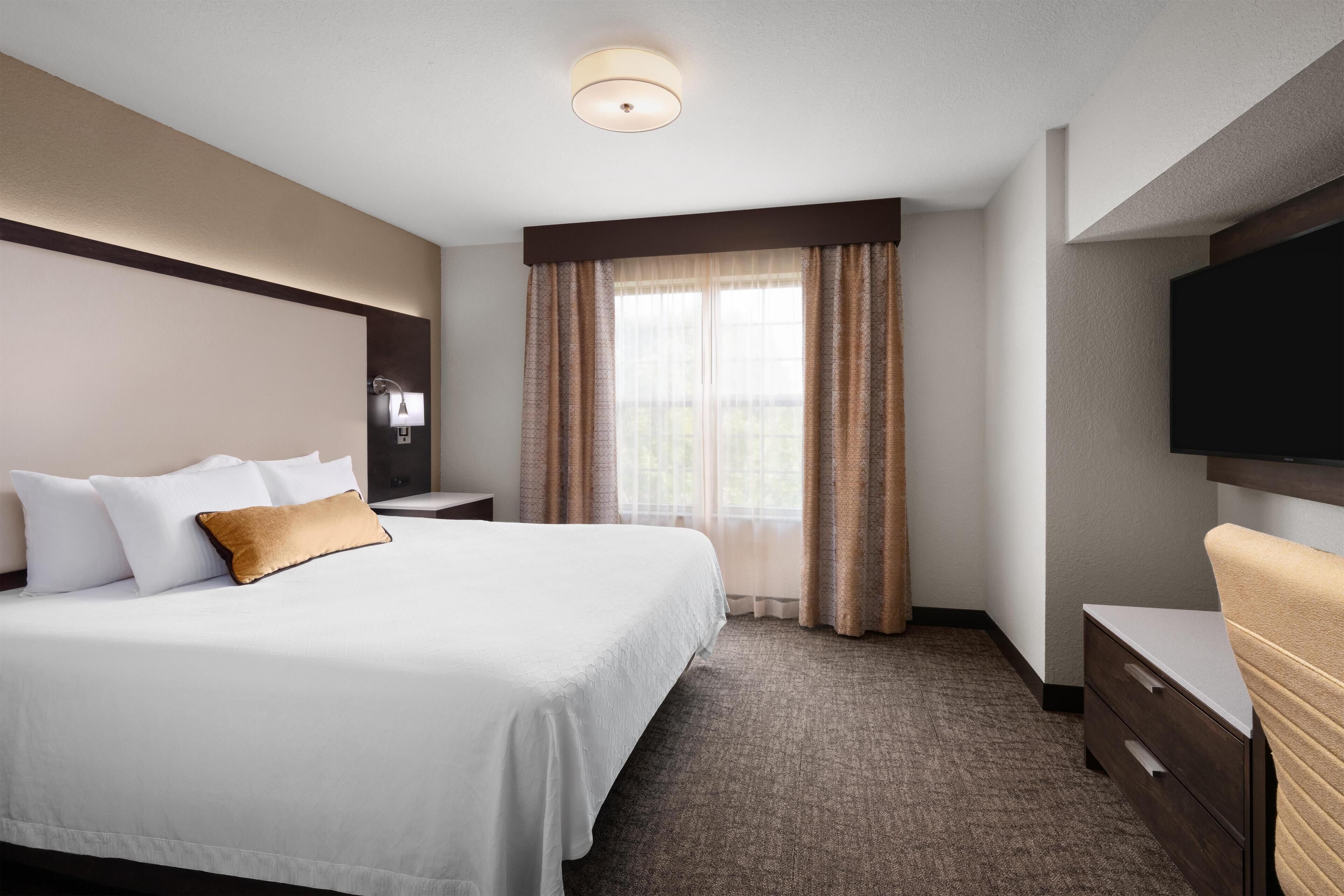 A hotel room with a large bed, a bedside table, a TV mounted on the wall, and a window with curtains.
