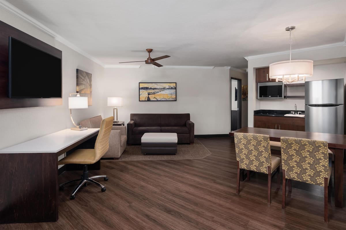 A hotel room with a desk, a sofa, a dining table with chairs, a ceiling fan, a TV, a microwave, and a refrigerator.