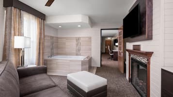 A modern hotel room with a sofa, ottoman, ceiling fan, large TV, fireplace, and a view into the hallway.
