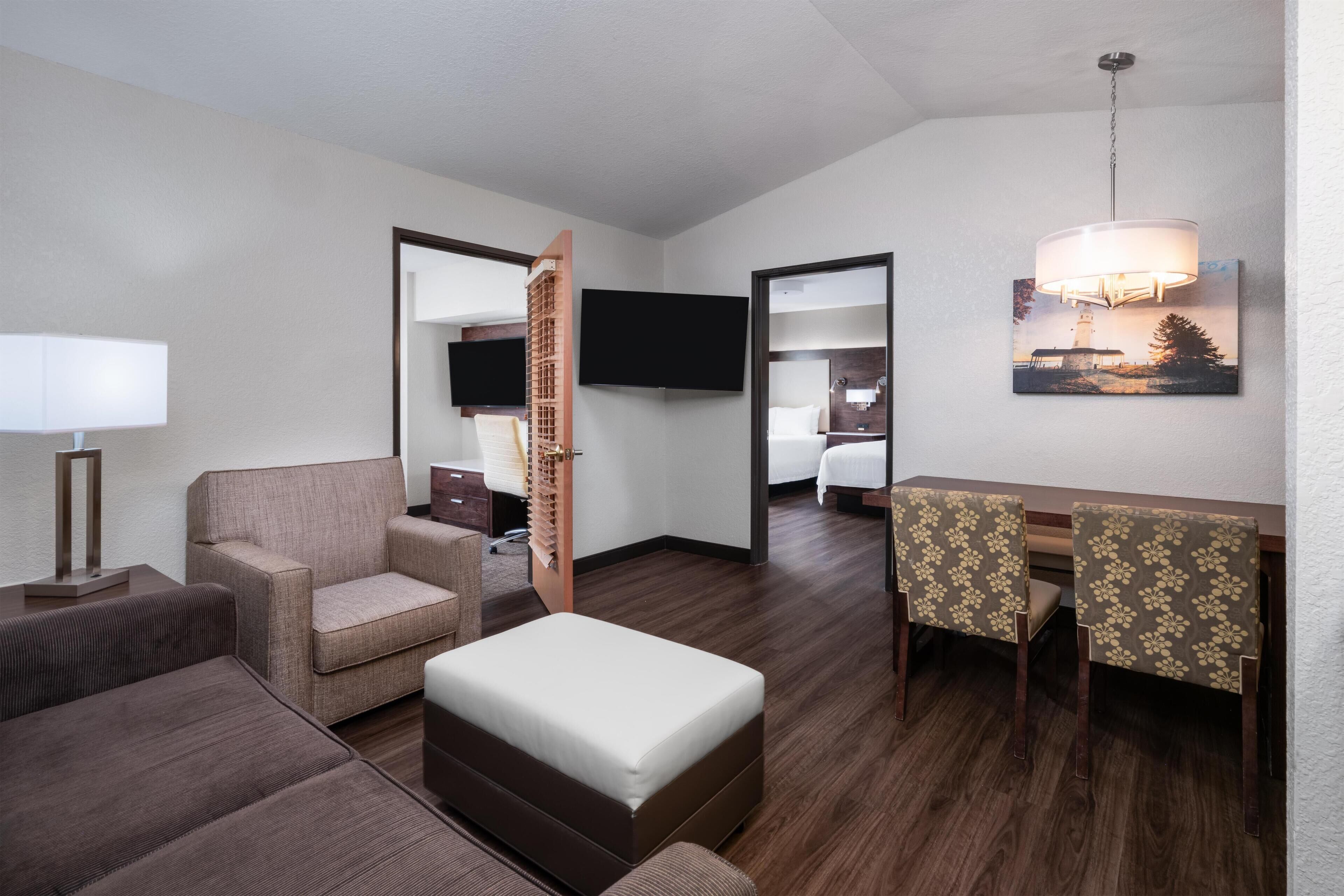 A hotel room with a sofa, ottoman, dining table with chairs, a TV, and a painting on the wall.