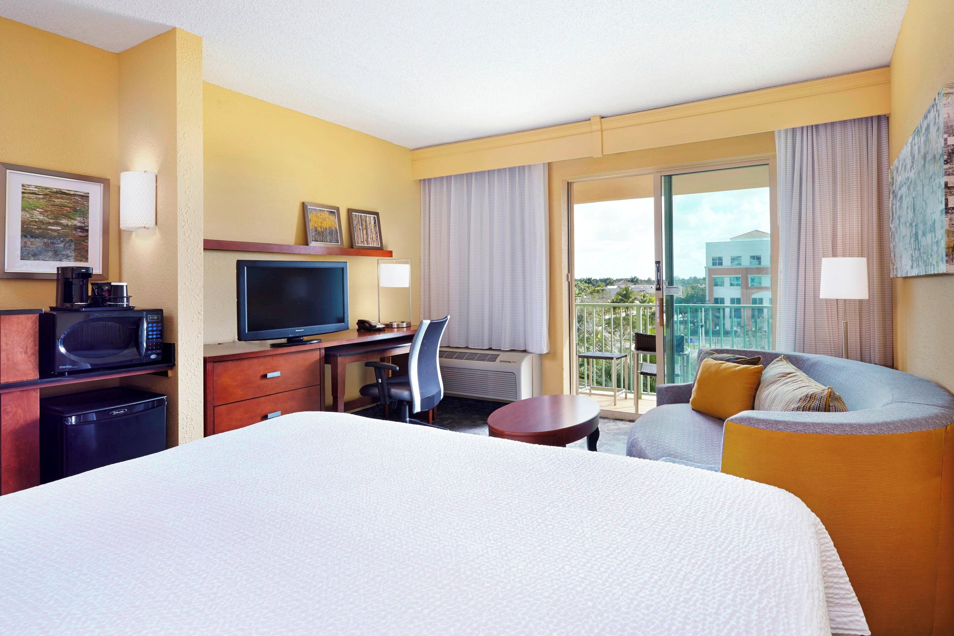 Ein Hotelzimmer mit einem großen Bett, einem Schreibtisch mit Fernseher, einem Sessel, einem kleinen Tisch und einem Balkon mit Blick auf Gebäude.