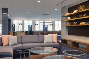 Un moderno vestíbulo de hotel con un sofá gris curvo, mesas de centro de vidrio y una estantería de madera con elementos decorativos.