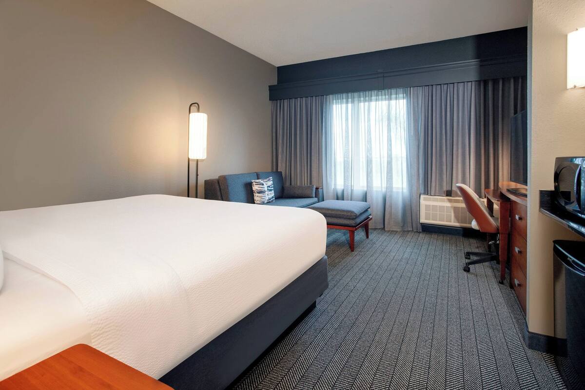 A hotel room with a large bed, a desk, a chair, a lamp, and a window with curtains.