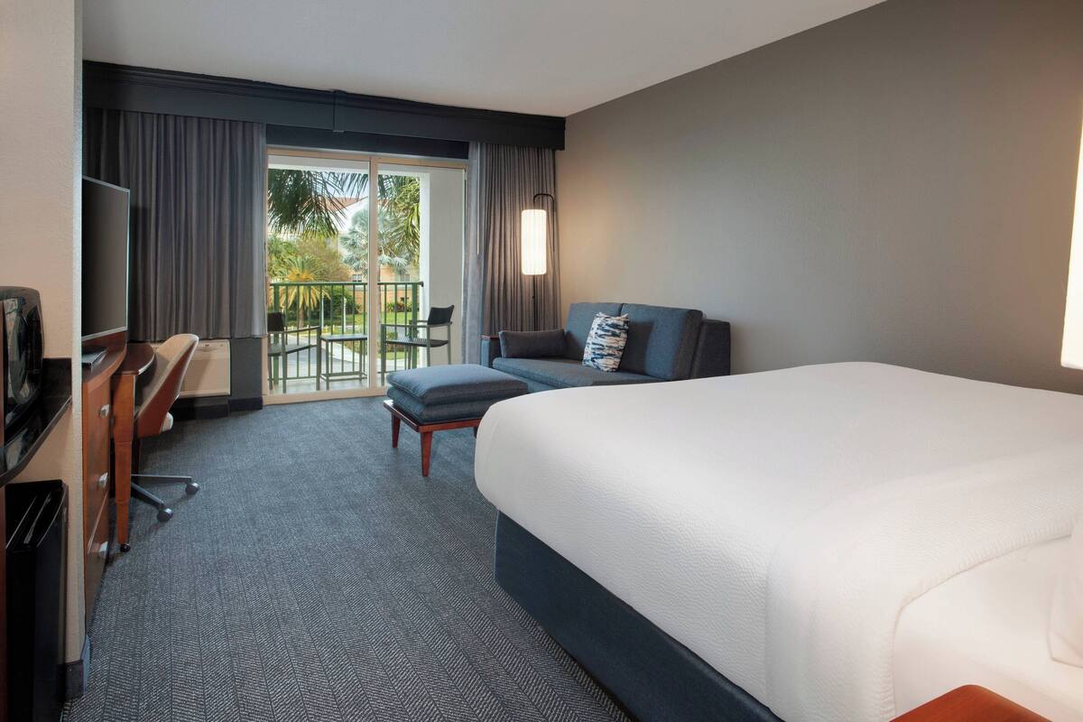 A hotel room with a large bed, a desk with a chair, a TV, and a balcony with curtains.