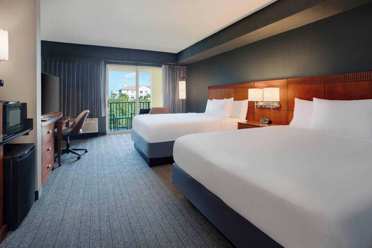 A hotel room with two beds, a desk with a chair, a television, and a balcony with a view of buildings.