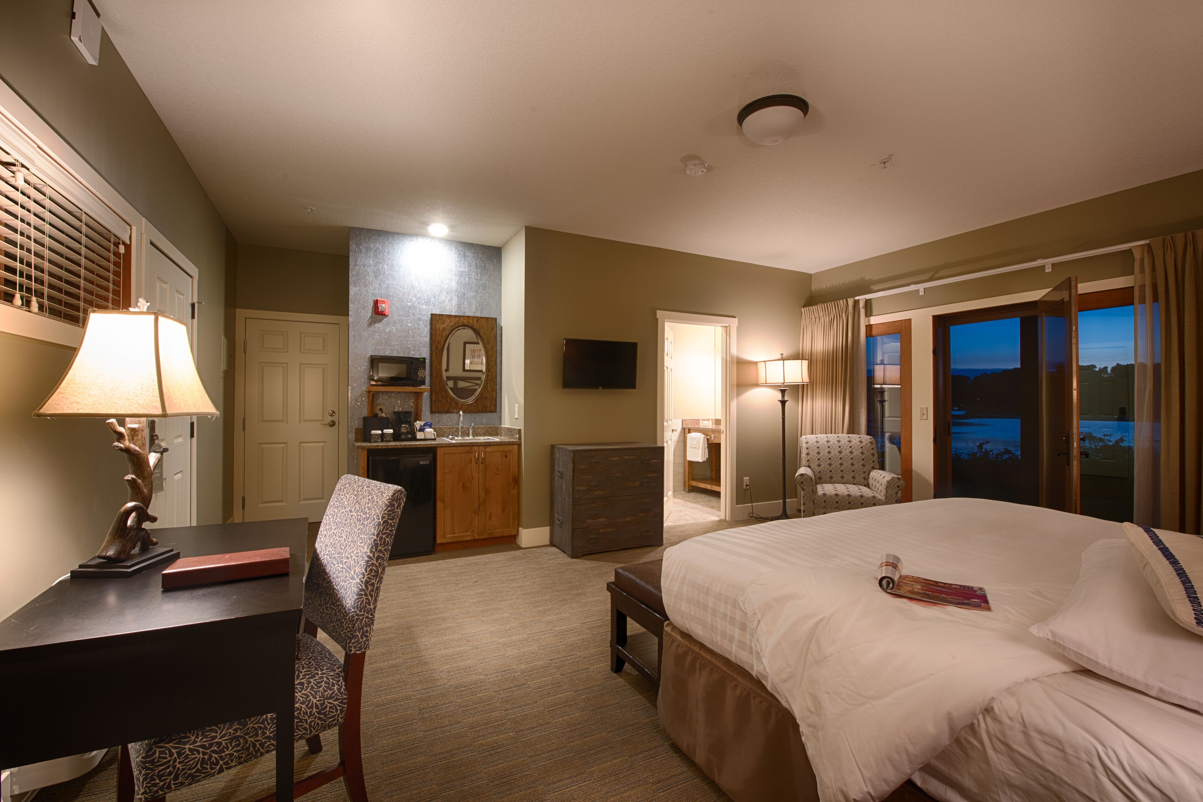 A hotel room with a bed, a desk, a chair, a kitchenette, and a view of the outdoors.