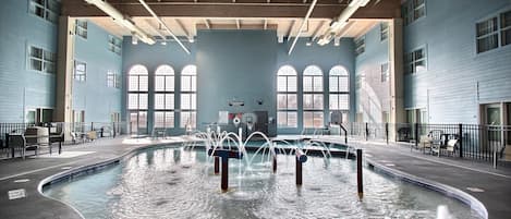 Una piscina interior con múltiples chorros de agua, un aro de baloncesto y un cartel de socorrista.