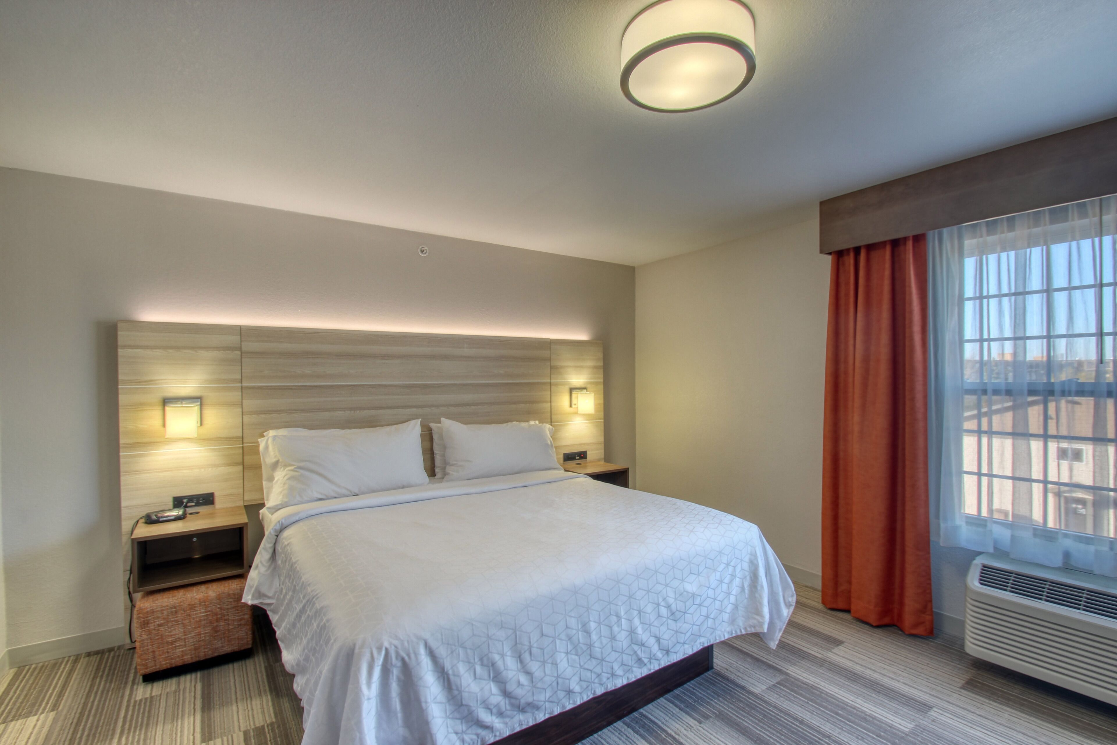 A hotel room with a large bed, bedside tables, a headboard with integrated lighting, a window with curtains, and a built-in air conditioning unit.