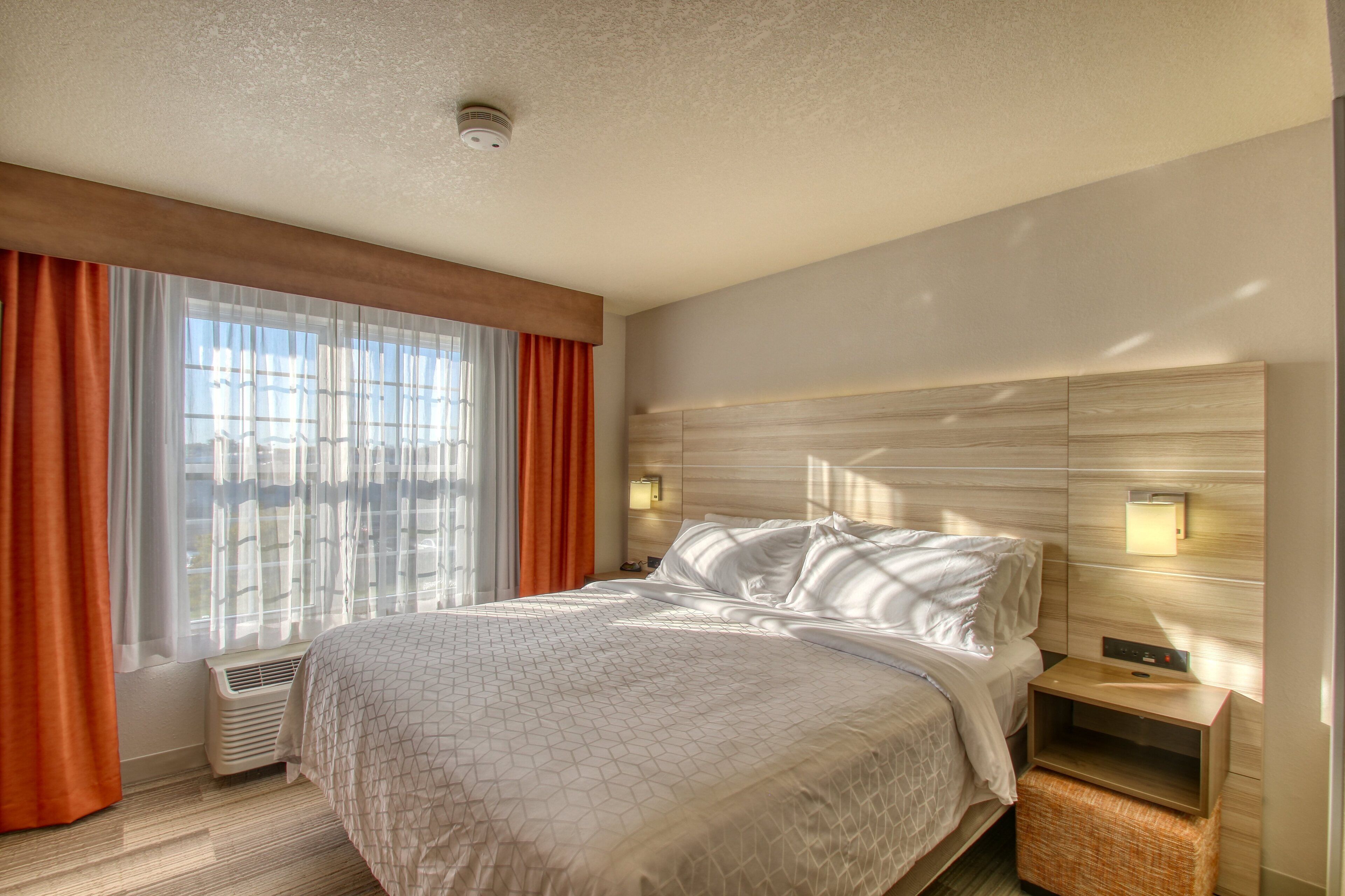 Een hotelkamer met een groot bed, een nachtkastje met een lamp, een raam met gordijnen en een wandradiator.