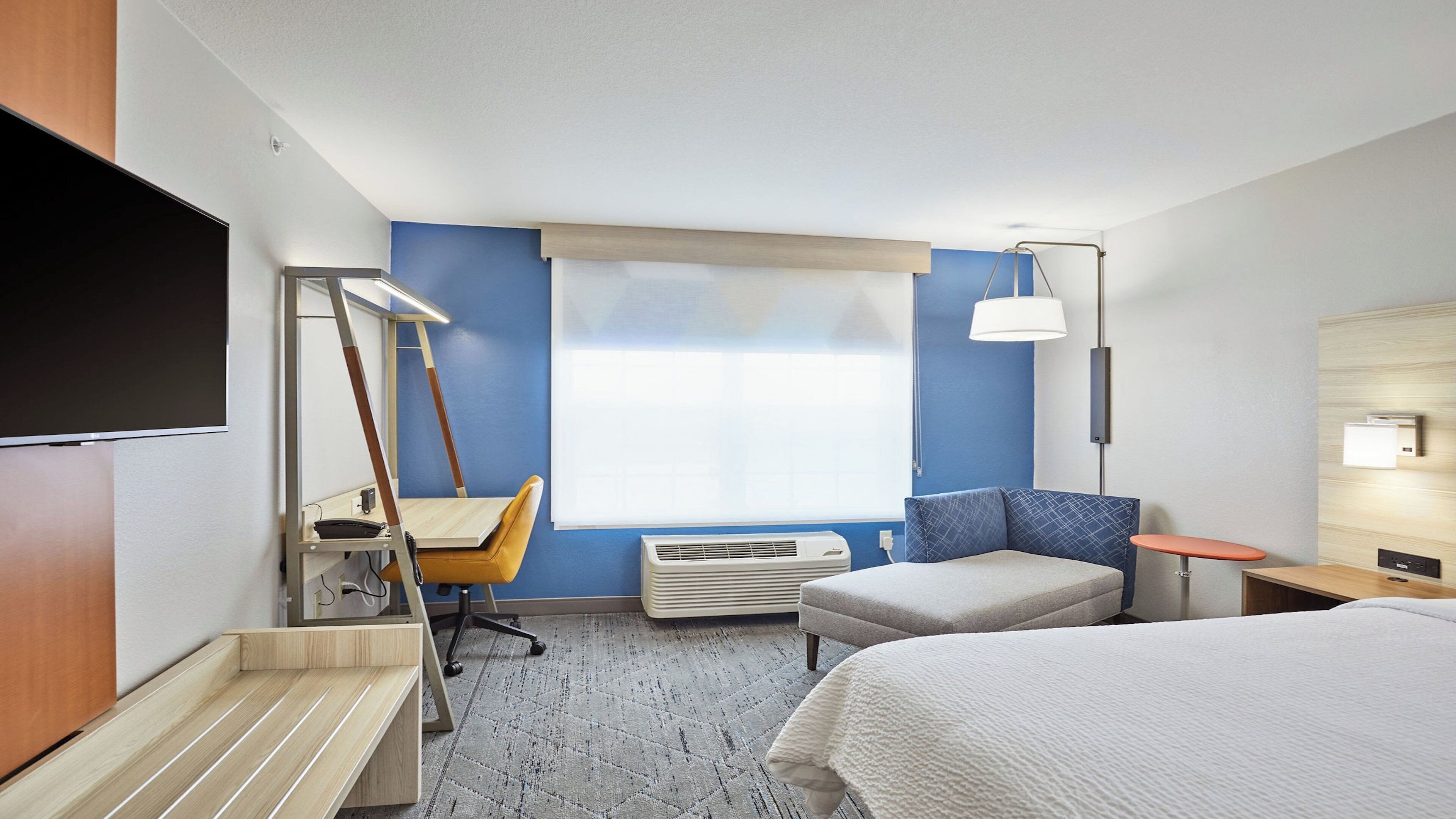 Habitación de hotel con una cama, un escritorio con silla, una lámpara de pie, un calentador y un televisor.