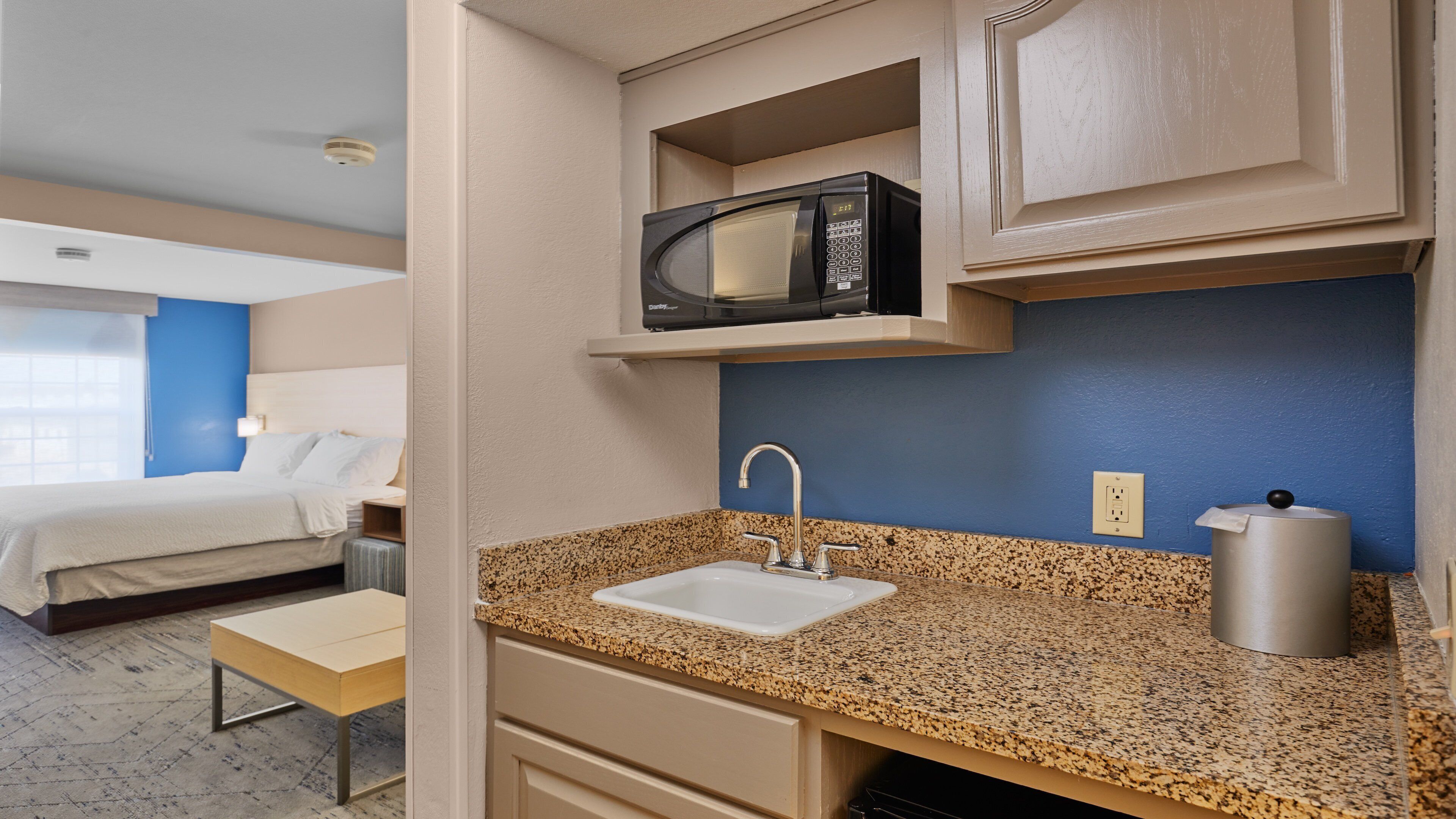 Una habitación de hotel con cama, kitchenette con microondas, lavamanos y una pequeña mesa.
