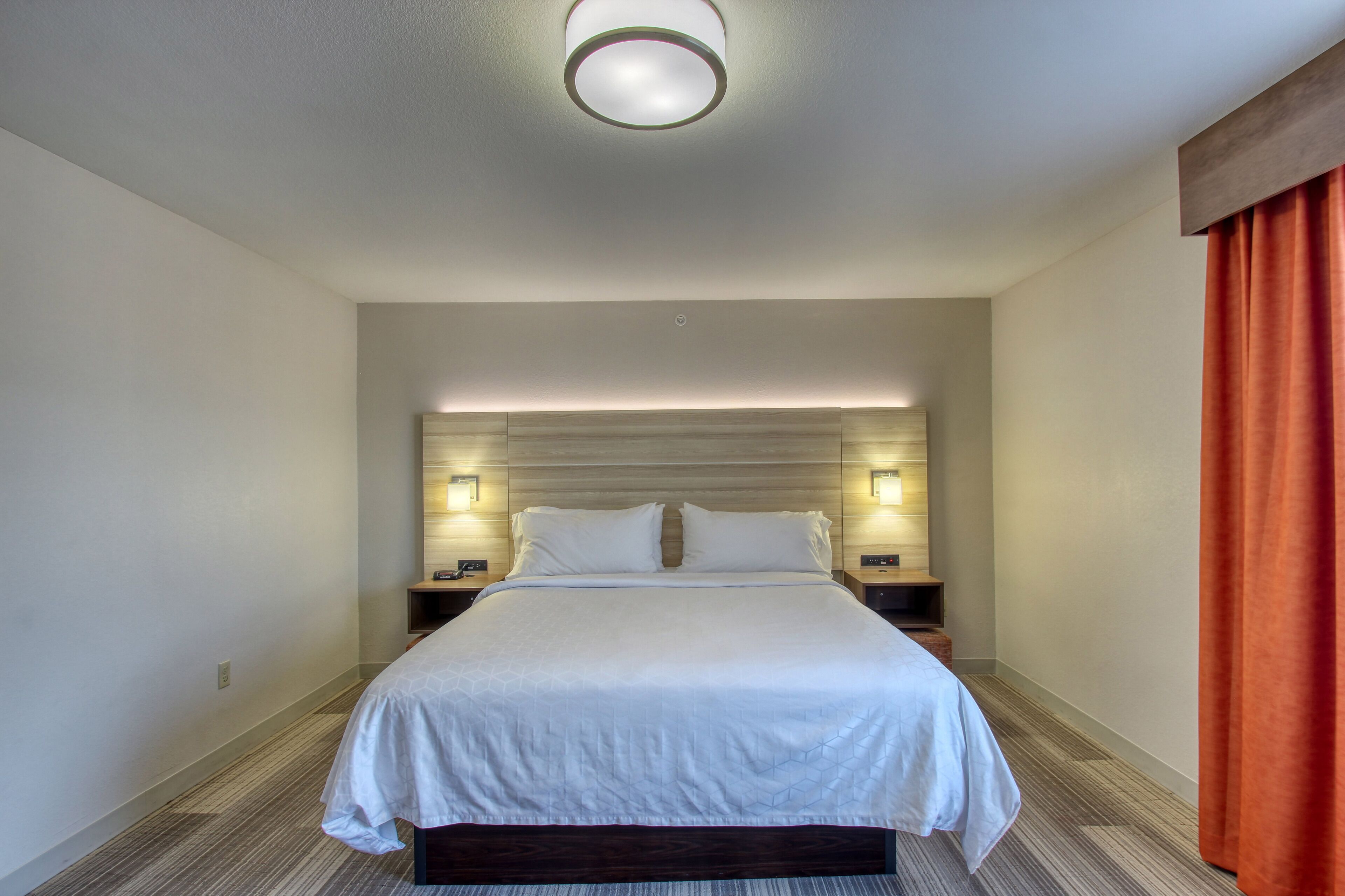 Habitación de hotel con una cama grande, dos mesitas de noche, cabecera con iluminación integrada y una cortina roja a la derecha.