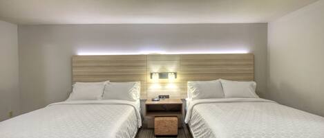 A hotel room with two beds, a wooden headboard, a nightstand with a lamp, and a small ottoman.