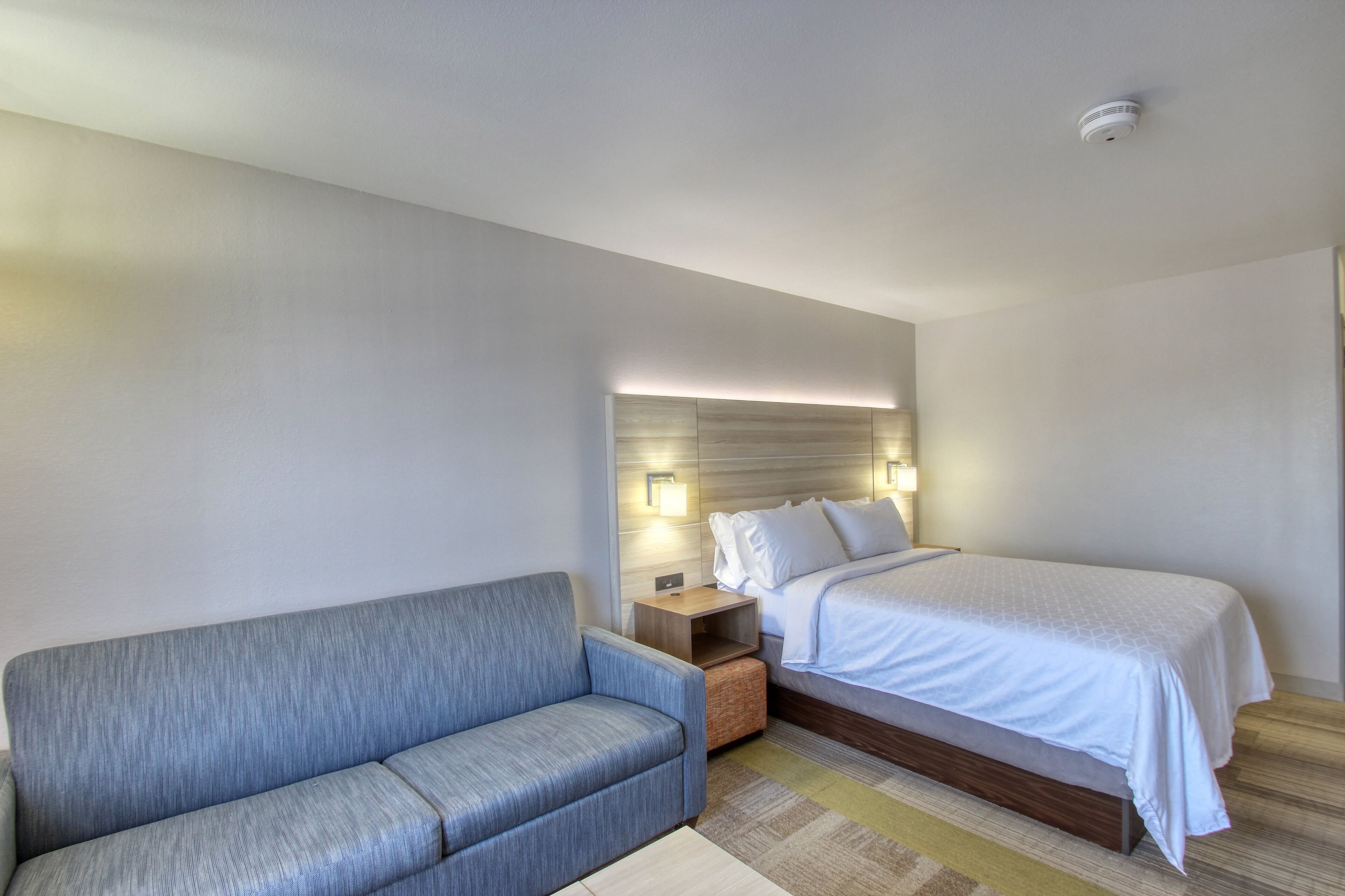 A hotel room with a bed, a sofa, a nightstand, and wall-mounted lights.