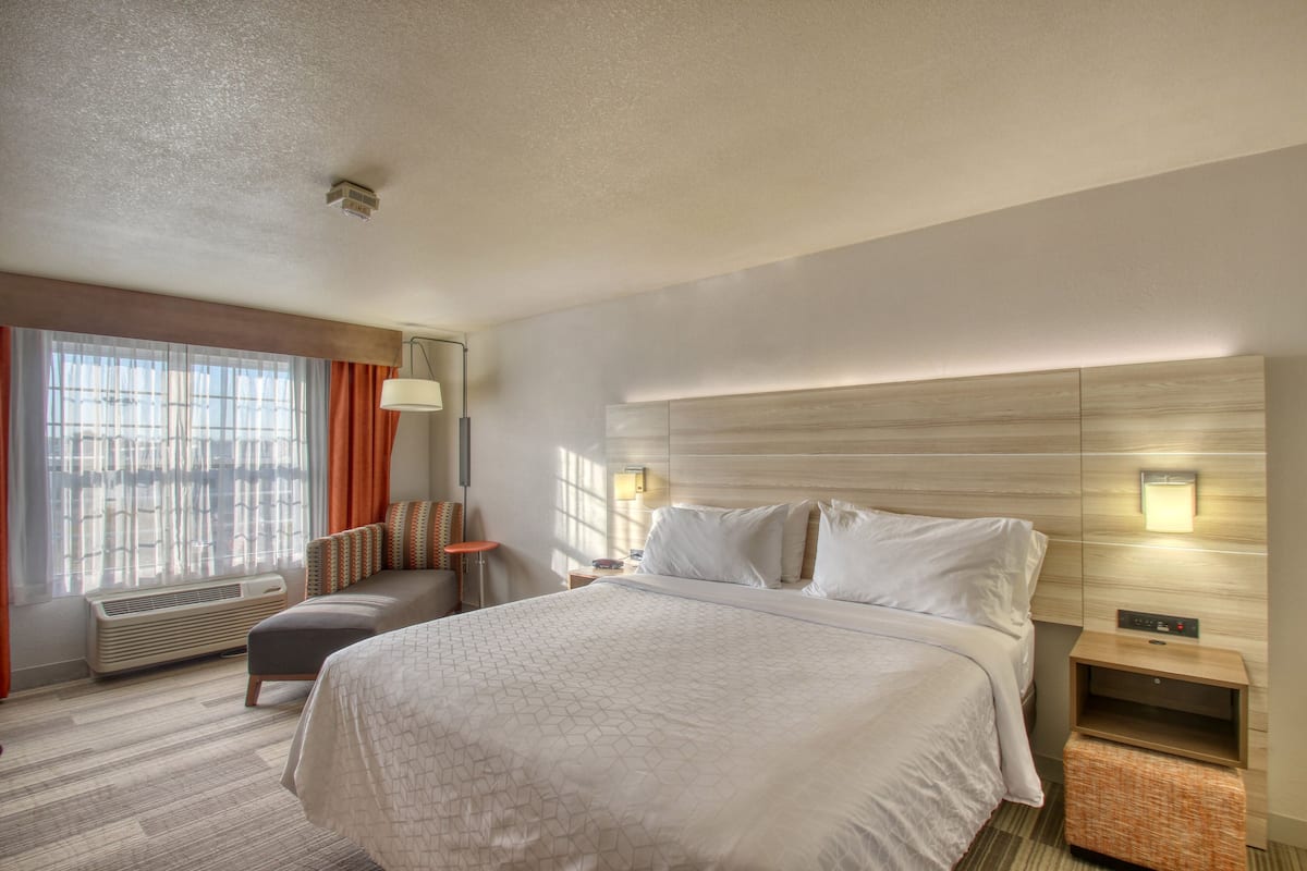 Ein Hotelzimmer mit einem großen Bett, einem Nachttisch, einer Lampe, einem Fenster mit Vorhängen und einer Wandleuchte.