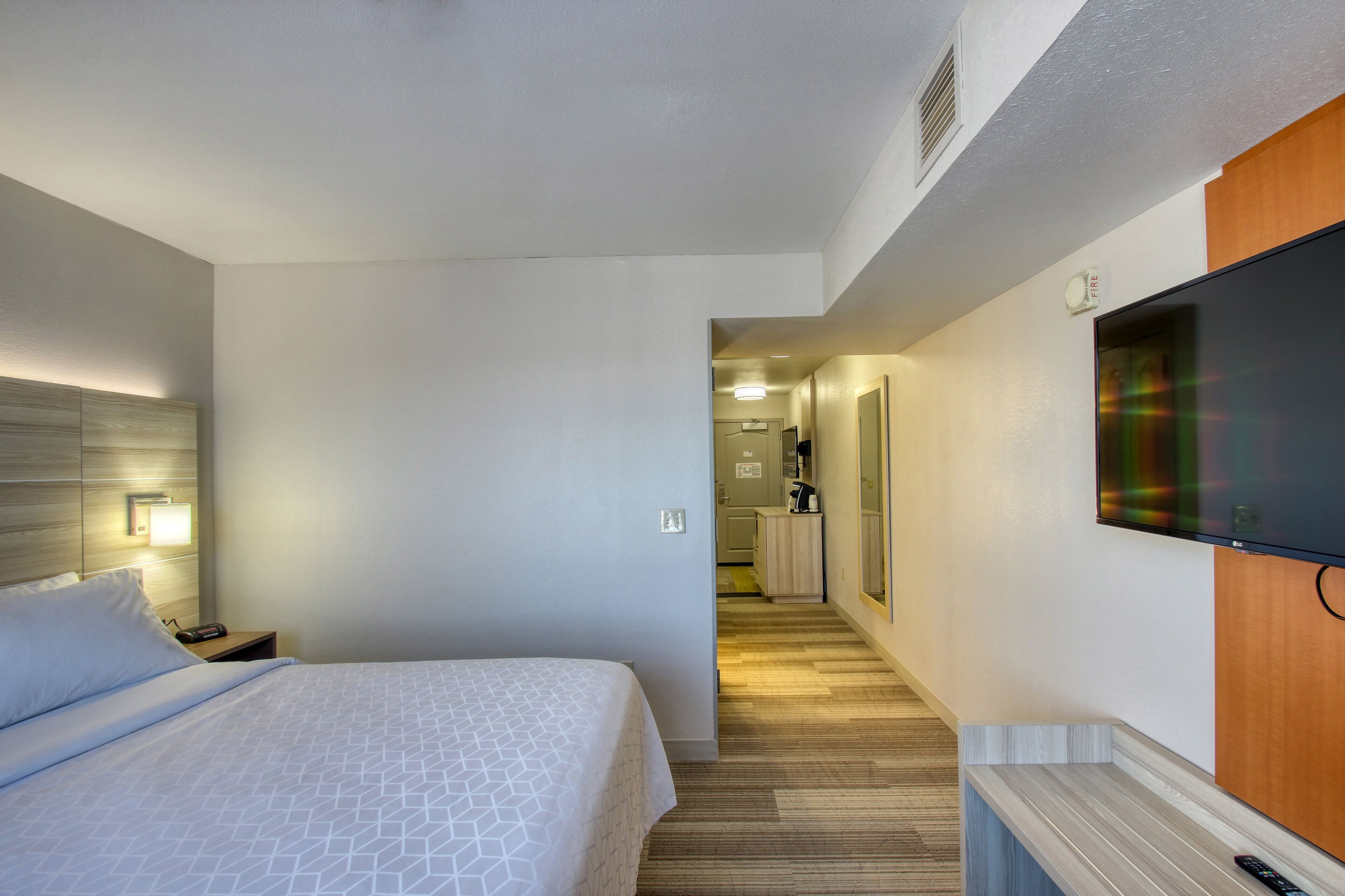 A hotel room with a bed, bedside table, TV, and a hallway leading to a bathroom.