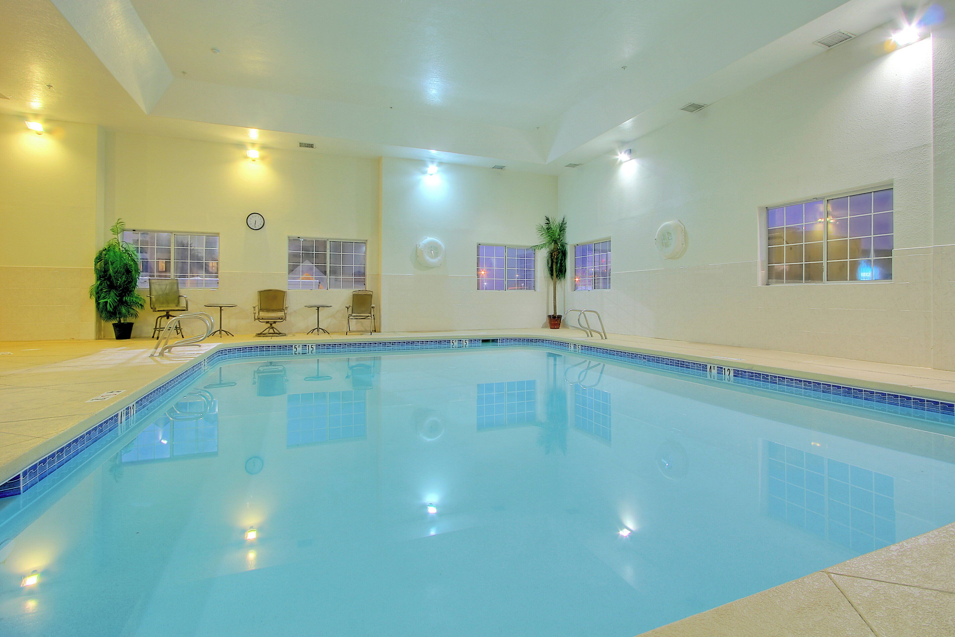 A swimming pool with clear water, surrounded by walls with windows, and equipped with chairs and potted plants.