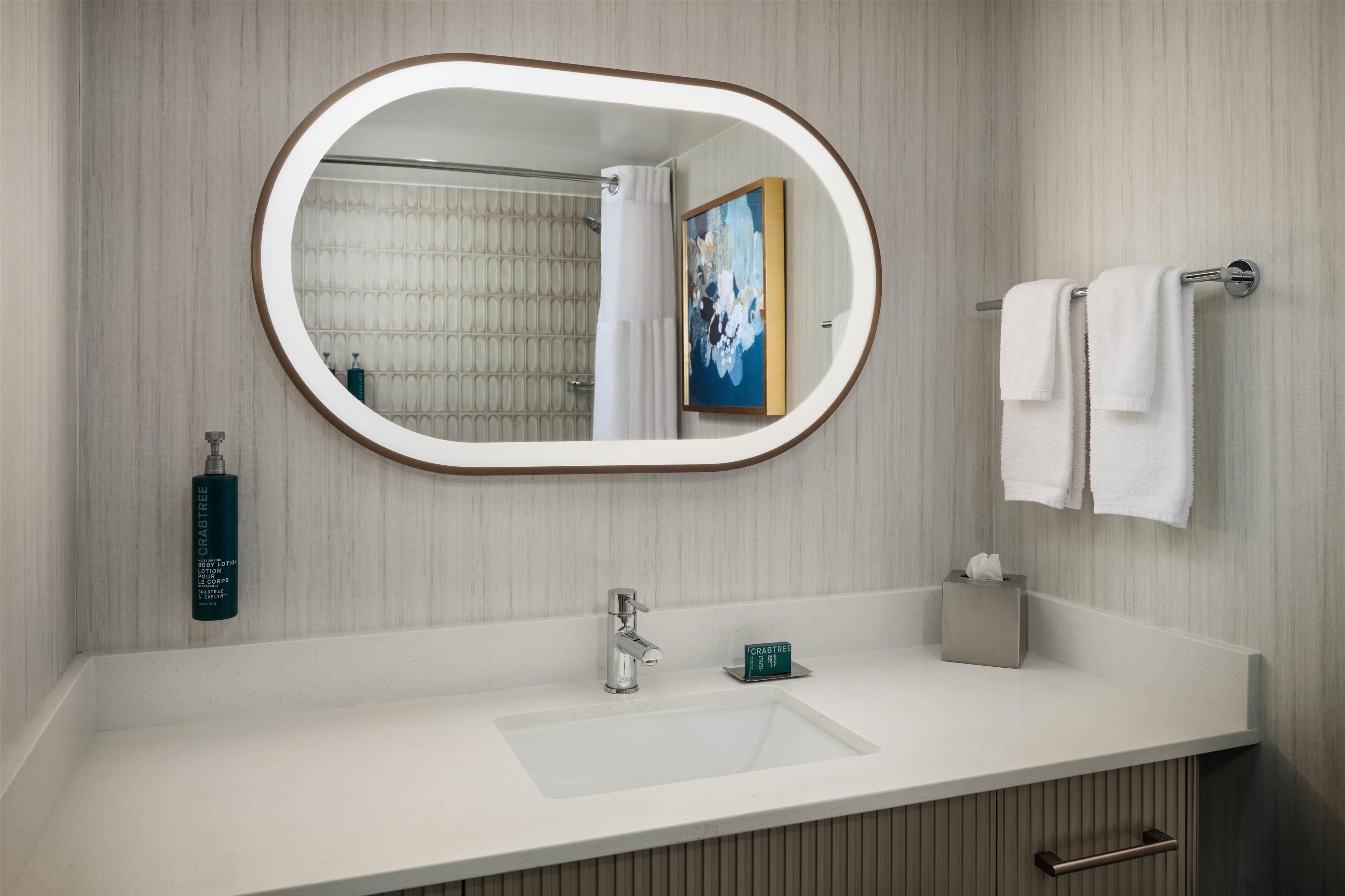 A bathroom with a white sink, a mirror with a light frame, a towel rack with two towels, a bottle of Crabtree lotion, and a painting on the wall.