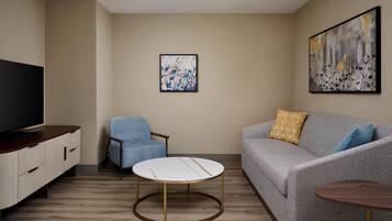 A modern living room with a grey sofa, a round coffee table, a blue armchair, a TV on a wooden cabinet, and abstract art on the walls.