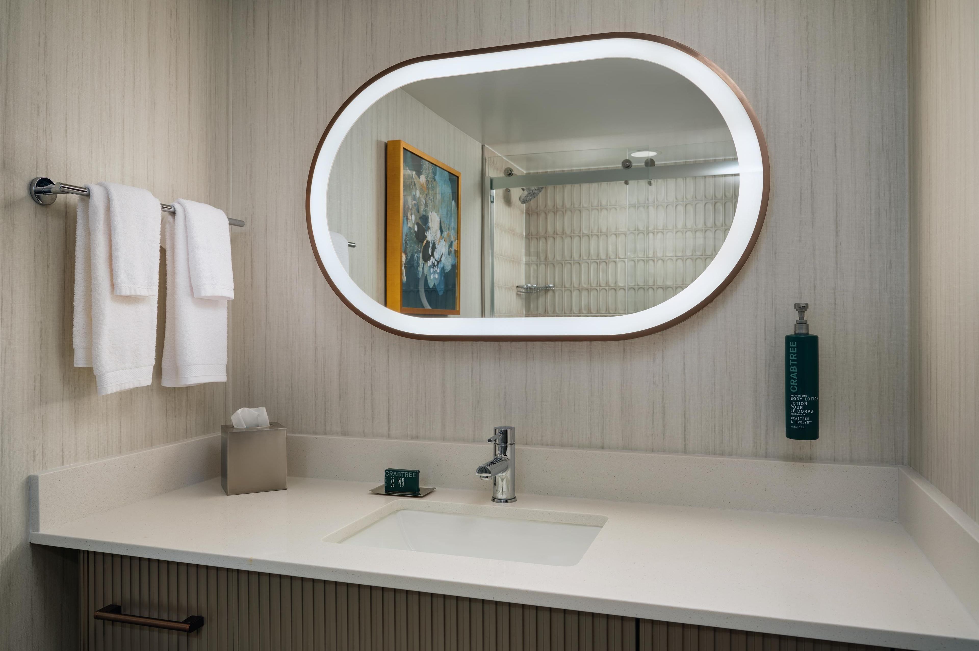 A bathroom with a white sink, a mirror, a towel rack with two white towels, a framed artwork, and a bottle of hand soap.